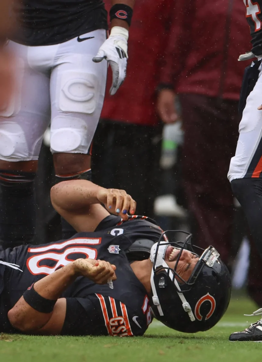 Caleb Williams - Ein Footballspieler im Trikot der Chicago Bears liegt auf dem Spielfeld und hält sich den Arm, während seine Teamkollegen während eines Spiels daneben stehen, nachdem die Bears Williams für verletzt erklärt haben. Alt-Text wurde KI-generiert.