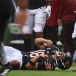 Caleb Williams - Ein Footballspieler im Trikot der Chicago Bears liegt auf dem Spielfeld und hält sich den Arm, während seine Teamkollegen während eines Spiels daneben stehen, nachdem die Bears Williams für verletzt erklärt haben. Alt-Text wurde KI-generiert.