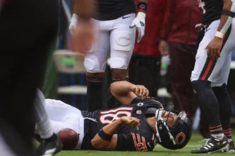 Caleb Williams - Ein Footballspieler im Trikot der Chicago Bears liegt auf dem Spielfeld und hält sich den Arm, während seine Teamkollegen während eines Spiels daneben stehen, nachdem die Bears Williams für verletzt erklärt haben. Alt-Text wurde KI-generiert.