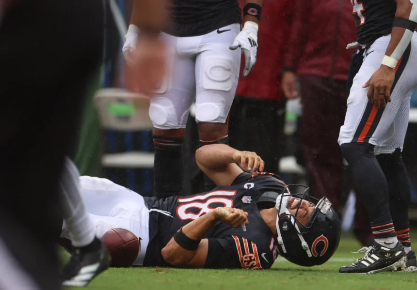 Caleb Williams - Ein Footballspieler im Trikot der Chicago Bears liegt auf dem Spielfeld und hält sich den Arm, während seine Teamkollegen während eines Spiels daneben stehen, nachdem die Bears Williams für verletzt erklärt haben. Alt-Text wurde KI-generiert.
