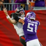 Baker Mayfield - Ein Footballspieler in einem violetten Trikot mit der Nummer 51 tackelt während eines Spiels einen Quarterback – möglicherweise Mayfield – in einem weißen Trikot. Während Mayfield spät zu Boden geht und auf den Zusammenprall nahe der Seitenlinie reagiert, verkörpern beide Spieler die Intensität dieses Duells der Bucs. Alt-Text wurde KI-generiert.