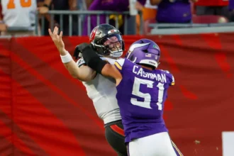 Baker Mayfield - Ein Footballspieler in einem violetten Trikot mit der Nummer 51 tackelt während eines Spiels einen Quarterback – möglicherweise Mayfield – in einem weißen Trikot. Während Mayfield spät zu Boden geht und auf den Zusammenprall nahe der Seitenlinie reagiert, verkörpern beide Spieler die Intensität dieses Duells der Bucs. Alt-Text wurde KI-generiert.