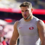 Nick Bosa - Ein muskulöser Footballspieler in einem ärmellosen Trikot der San Francisco 49ers steht auf einem Spielfeld, im Hintergrund sind verschwommene Stadionplätze zu sehen – bereit, im Saisonauftakt der 49ers zu dominieren, wenn Bosa spielt. Alt-Text wurde KI-generiert.