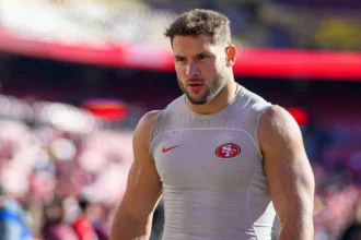 Nick Bosa - Ein muskulöser Footballspieler in einem ärmellosen Trikot der San Francisco 49ers steht auf einem Spielfeld, im Hintergrund sind verschwommene Stadionplätze zu sehen – bereit, im Saisonauftakt der 49ers zu dominieren, wenn Bosa spielt. Alt-Text wurde KI-generiert.