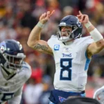 Will Levis - Der Quarterback der Tennessee Titans im weißen Trikot hebt beide Hände, während er während eines Footballspiels einen Spielzug ansagt; im Hintergrund sind Teamkollegen und Gegner zu sehen – darunter die Jets, die zwei Quarterbacks einsetzen. Alt-Text wurde KI-generiert.