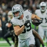 Ein Footballspieler in einem silber-schwarzen Trikot, das an die Raiders erinnert, läuft während eines Spiels mit dem Ball, im Hintergrund sind Mitspieler und Gegner zu sehen. Laut Bericht fällt Brock Bowers wegen einer Verletzung aus. Alt-Text wurde KI-generiert.