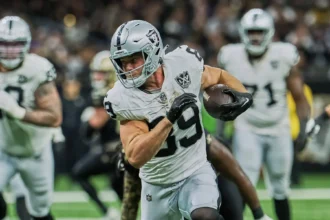 Ein Footballspieler in einem silber-schwarzen Trikot, das an die Raiders erinnert, läuft während eines Spiels mit dem Ball, im Hintergrund sind Mitspieler und Gegner zu sehen. Laut Bericht fällt Brock Bowers wegen einer Verletzung aus. Alt-Text wurde KI-generiert.