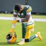 Jayden Reed - Ein Footballspieler im Trikot der Packers, der als Jayden Reed identifiziert wurde, kniet auf einem Knie auf dem Spielfeld, hält seinen Helm in einer Hand und scheint vor dem nächsten Spielzug eine Pause einzulegen und sich zu konzentrieren. Alt-Text wurde KI-generiert.