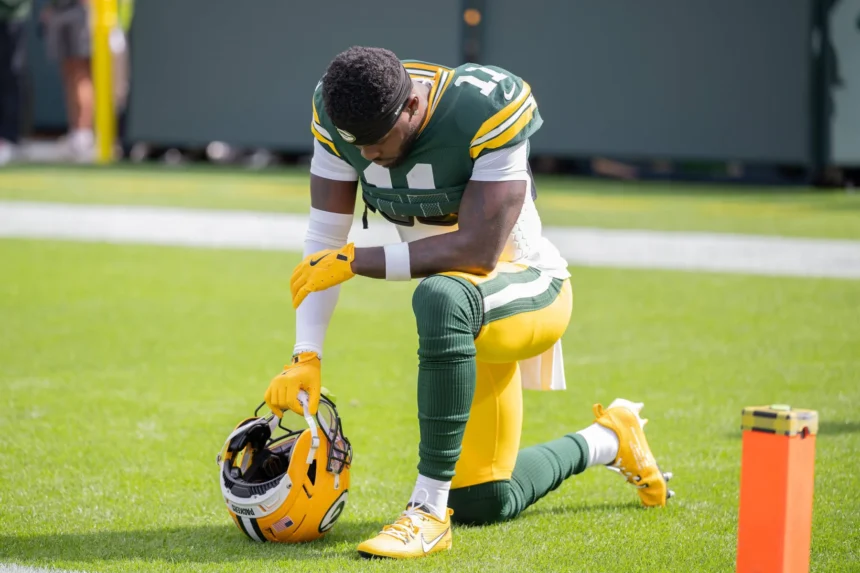 Jayden Reed - Ein Footballspieler im Trikot der Packers, der als Jayden Reed identifiziert wurde, kniet auf einem Knie auf dem Spielfeld, hält seinen Helm in einer Hand und scheint vor dem nächsten Spielzug eine Pause einzulegen und sich zu konzentrieren. Alt-Text wurde KI-generiert.
