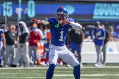 Malik Nabers - Ein Footballspieler in blauer Spielkleidung steht mitten in einer Jubelszene auf dem Spielfeld und hält einen Football in der Hand; im Hintergrund sind Mannschaftspersonal und die Anzeigetafel zu sehen, während sich die Giants auf Nabers für den Sonntagabend vorbereiten. Alt-Text wurde KI-generiert.