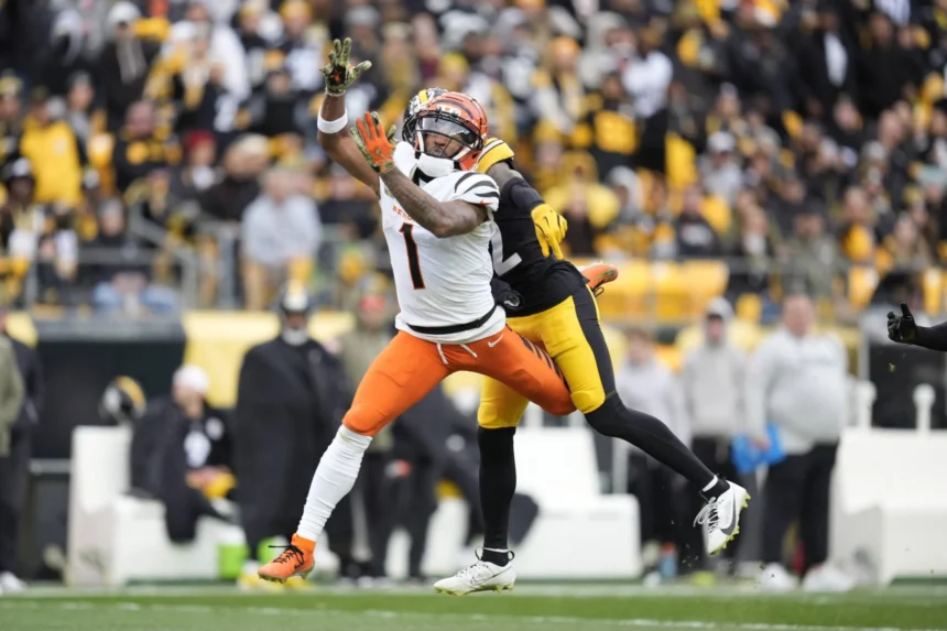 Ein Footballspieler in einem weiß-orangefarbenen Trikot versucht, einen Pass zu fangen, während er auf dem Spielfeld während eines NFL-Spiels von einem Spieler in einem schwarz-gelben Trikot eng gedeckt wird. Dabei können „Jubelstrafen“ – also Strafen für Jubelgesten – dem Geschehen zusätzliche Dramatik verleihen. Alt-Text wurde KI-generiert.