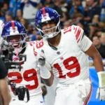 Jameis Winston - Footballspieler der New York Giants in weißen Trikots feiern während eines Spiels auf dem Feld; ein Spieler mit der Rückennummer 19 lächelt und gestikuliert, während die Schlagzeilen melden: „Bericht: Giants und Winston einigen sich“. Alt-Text wurde KI-generiert.