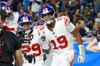 Jameis Winston - Footballspieler der New York Giants in weißen Trikots feiern während eines Spiels auf dem Feld; ein Spieler mit der Rückennummer 19 lächelt und gestikuliert, während die Schlagzeilen melden: „Bericht: Giants und Winston einigen sich“. Alt-Text wurde KI-generiert.