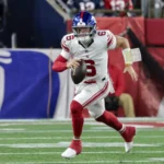 Jaxson Dart - Ein Spieler der Giants in einem weiß-roten Trikot sprintet während eines Spiels auf einem grünen Rasen mit dem Ball los. Alt-Text wurde KI-generiert.