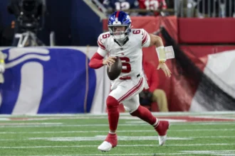Jaxson Dart - Ein Spieler der Giants in einem weiß-roten Trikot sprintet während eines Spiels auf einem grünen Rasen mit dem Ball los. Alt-Text wurde KI-generiert.