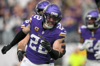 Ein Footballspieler in einem violetten Trikot und Helm der Minnesota Vikings läuft während eines Spiels über das Spielfeld, im Hintergrund sind seine Teamkollegen zu sehen, während der Bericht „Vikings holen Harrison Smith“ die starke Defensivaufstellung des Teams unterstreicht. Alt-Text wurde KI-generiert.