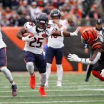 Kyle Monangai - Ein Footballspieler der „Bears“ in einem weißen Trikot läuft mit dem Ball über das Spielfeld und weicht dabei einem Verteidiger in schwarz-orangefarbener Spielkleidung aus. Alt-Text wurde KI-generiert.