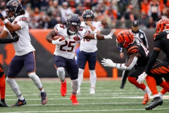 Kyle Monangai - Ein Footballspieler der „Bears“ in einem weißen Trikot läuft mit dem Ball über das Spielfeld und weicht dabei einem Verteidiger in schwarz-orangefarbener Spielkleidung aus. Alt-Text wurde KI-generiert.