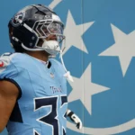Cedric Gray - Ein Footballspieler der Tennessee Titans in voller Spielkleidung steht vor einer blauen Wand mit drei weißen Sternen und präsentiert dabei die kräftigen Farben des „Titans Gray nach UTV“-Alternativdesigns. Alt-Text wurde KI-generiert.