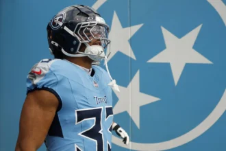 Cedric Gray - Ein Footballspieler der Tennessee Titans in voller Spielkleidung steht vor einer blauen Wand mit drei weißen Sternen und präsentiert dabei die kräftigen Farben des „Titans Gray nach UTV“-Alternativdesigns. Alt-Text wurde KI-generiert.