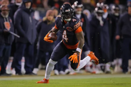 D’Andre Swift - Ein Footballspieler in einem dunklen Trikot rennt während eines Spiels mit dem Ball über das Spielfeld, während im Hintergrund andere Spieler und Trainer zu sehen sind – darunter auch Mitglieder der Bericht Bears, die gerade ihren Vertrag mit Swift verlängern. Alt-Text wurde KI-generiert.