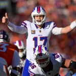 Josh Allen - Ein Footballspieler in einem weißen Trikot mit der Nummer 17 reckt auf dem Spielfeld beide Daumen in die Höhe, umgeben von Mannschaftskameraden und gegnerischen Spielern in orangefarbenen Trikots. Alt-Text wurde KI-generiert.