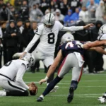 Patriots-Trick - Ein Football-Kicker versucht während eines Spiels ein Field Goal, während ein Mitspieler den Ball hält und gegnerische Spieler auf sie zustürmen; gleichzeitig legt die NFL Regeln für Videowände fest, um den fairen Spielablauf zu fördern und die Aufmerksamkeit auf das Spielgeschehen zu lenken. Alt-Text wurde KI-generiert.