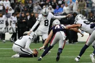Patriots-Trick - Ein Football-Kicker versucht während eines Spiels ein Field Goal, während ein Mitspieler den Ball hält und gegnerische Spieler auf sie zustürmen; gleichzeitig legt die NFL Regeln für Videowände fest, um den fairen Spielablauf zu fördern und die Aufmerksamkeit auf das Spielgeschehen zu lenken. Alt-Text wurde KI-generiert.