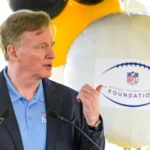 Roger Goodell - Ein Mann in einem blauen Hemd und einem dunklen Blazer spricht ins Mikrofon und gestikuliert dabei mit der Hand; hinter ihm ist ein Luftballon mit dem Logo der NFL Foundation zu sehen, der an die jüngsten Ereignisse erinnert, bei denen Goodell eine Vertragsverlängerung unterzeichnet hat. Alt-Text wurde KI-generiert.