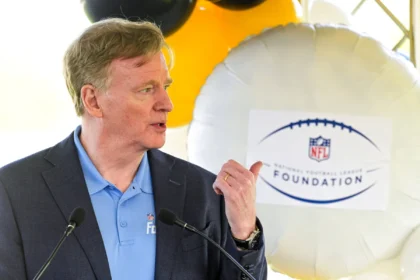 Roger Goodell - Ein Mann in einem blauen Hemd und einem dunklen Blazer spricht ins Mikrofon und gestikuliert dabei mit der Hand; hinter ihm ist ein Luftballon mit dem Logo der NFL Foundation zu sehen, der an die jüngsten Ereignisse erinnert, bei denen Goodell eine Vertragsverlängerung unterzeichnet hat. Alt-Text wurde KI-generiert.