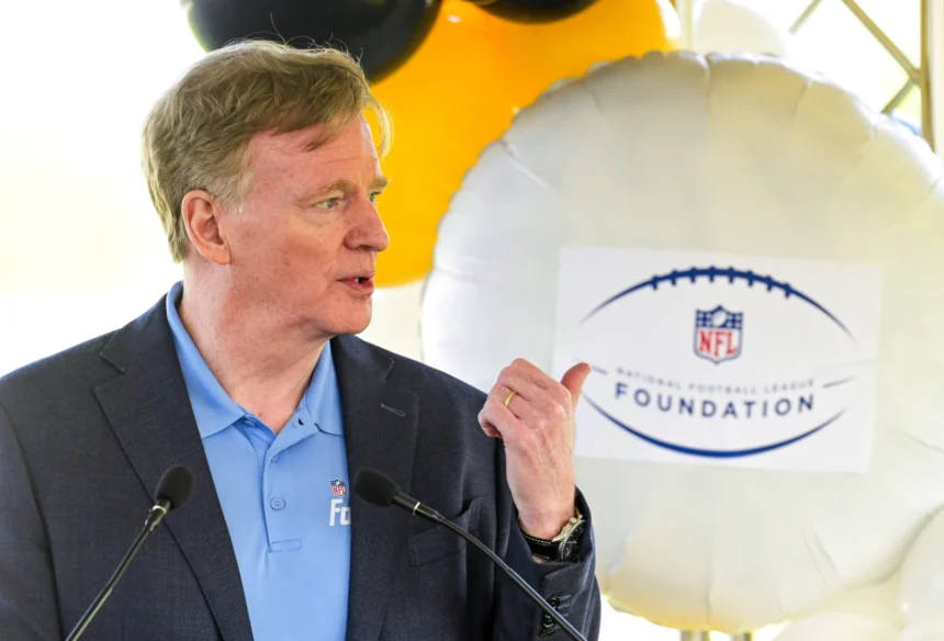 Roger Goodell - Ein Mann in einem blauen Hemd und einem dunklen Blazer spricht ins Mikrofon und gestikuliert dabei mit der Hand; hinter ihm ist ein Luftballon mit dem Logo der NFL Foundation zu sehen, der an die jüngsten Ereignisse erinnert, bei denen Goodell eine Vertragsverlängerung unterzeichnet hat. Alt-Text wurde KI-generiert.