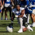 Myles Garrett kniet während des Trainings auf dem Spielfeld, hält einen Helm in der Hand und ist von Teamkollegen in blau-weißen Trikots umgeben, während Garrett sich gegen Allen behauptet und dabei Entschlossenheit und Teamgeist unter Beweis stellt. Alt-Text wurde KI-generiert.