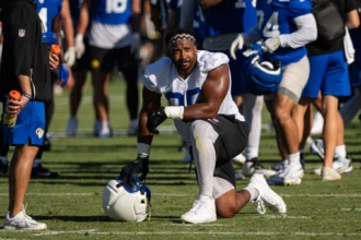 Myles Garrett kniet während des Trainings auf dem Spielfeld, hält einen Helm in der Hand und ist von Teamkollegen in blau-weißen Trikots umgeben, während Garrett sich gegen Allen behauptet und dabei Entschlossenheit und Teamgeist unter Beweis stellt. Alt-Text wurde KI-generiert.