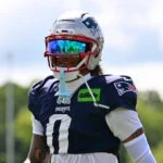 Christian Gonzalez - Ein Footballspieler in Trikot und Helm der New England Patriots steht während des Trainings im Freien, trägt ein getöntes Visier und hält einen Mundschutz in der Hand – kurz nachdem bekannt wurde, dass sich die Patriots und Gonzalez geeinigt haben. Alt-Text wurde KI-generiert.