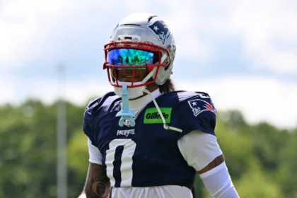 Christian Gonzalez - Ein Footballspieler in Trikot und Helm der New England Patriots steht während des Trainings im Freien, trägt ein getöntes Visier und hält einen Mundschutz in der Hand – kurz nachdem bekannt wurde, dass sich die Patriots und Gonzalez geeinigt haben. Alt-Text wurde KI-generiert.