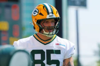 Tucker Kraft - Ein Footballspieler, der einen Helm und ein Trikot der Green Bay Packers mit der Nummer 85 trägt, steht bei Tageslicht auf dem Spielfeld, während einige Fans gespannt seine Leistung verfolgen. Alt-Text wurde KI-generiert.