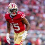 De'Zhaun Stribling - Ein Footballspieler in einem rot-goldenen Trikot mit der Nummer 15 bereitet sich auf einen Spielzug auf dem Feld vor – ganz im dynamischen Stil, für den Shanahan von den 49ers bekannt ist –, während im Hintergrund eine verschwommene Menschenmenge zu sehen ist. Alt-Text wurde KI-generiert.