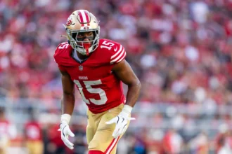 De'Zhaun Stribling - Ein Footballspieler in einem rot-goldenen Trikot mit der Nummer 15 bereitet sich auf einen Spielzug auf dem Feld vor – ganz im dynamischen Stil, für den Shanahan von den 49ers bekannt ist –, während im Hintergrund eine verschwommene Menschenmenge zu sehen ist. Alt-Text wurde KI-generiert.