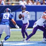 Ein Football-Quarterback in einem weiß-lila Trikot, das an die Vikings erinnert, macht sich bereit, den Ball zu werfen, während Verteidiger und Angreifer auf dem sonnigen Spielfeld näherkommen – genau so, wie J.J. McCarthy mit Entschlossenheit und Konzentration in die Saison gestartet ist. Alt-Text wurde KI-generiert.