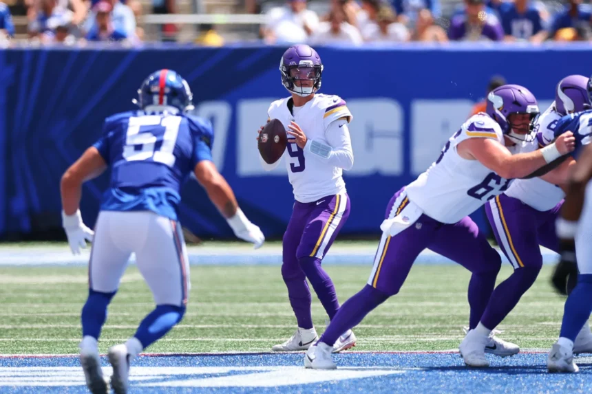 Ein Football-Quarterback in einem weiß-lila Trikot, das an die Vikings erinnert, macht sich bereit, den Ball zu werfen, während Verteidiger und Angreifer auf dem sonnigen Spielfeld näherkommen – genau so, wie J.J. McCarthy mit Entschlossenheit und Konzentration in die Saison gestartet ist. Alt-Text wurde KI-generiert.