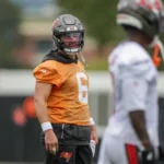 Baker Mayfield - Ein Footballspieler in einem orangefarbenen Trikot und Helm steht mit anderen Spielern in weißen Trikots auf einem Trainingsplatz und blickt nach links – was vielleicht an Bericht Mayfield und die Bucs während ihrer Trainingseinheiten erinnert. Alt-Text wurde KI-generiert.