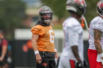 Baker Mayfield - Ein Footballspieler in einem orangefarbenen Trikot und Helm steht mit anderen Spielern in weißen Trikots auf einem Trainingsplatz und blickt nach links – was vielleicht an Bericht Mayfield und die Bucs während ihrer Trainingseinheiten erinnert. Alt-Text wurde KI-generiert.