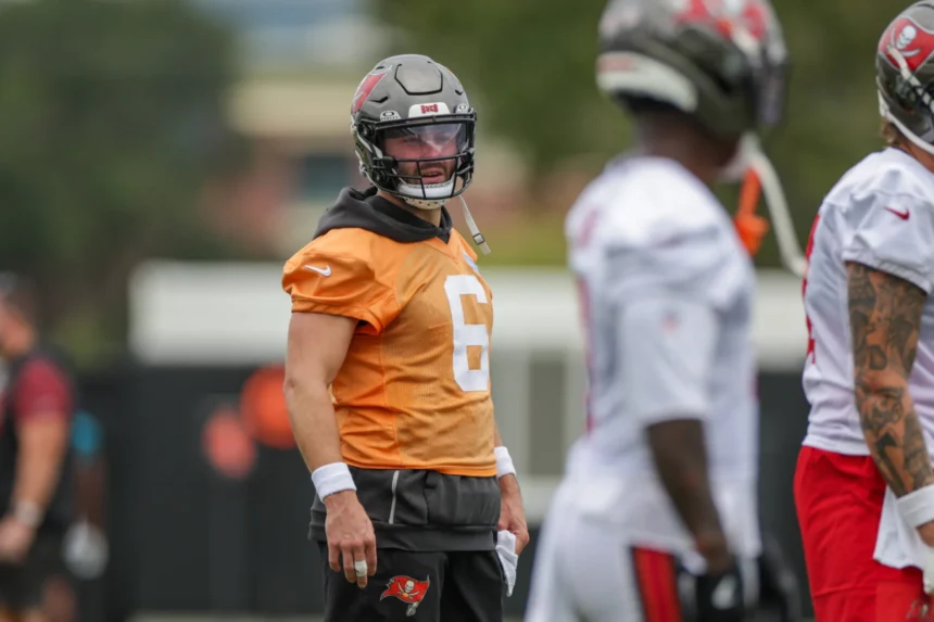 Baker Mayfield - Ein Footballspieler in einem orangefarbenen Trikot und Helm steht mit anderen Spielern in weißen Trikots auf einem Trainingsplatz und blickt nach links – was vielleicht an Bericht Mayfield und die Bucs während ihrer Trainingseinheiten erinnert. Alt-Text wurde KI-generiert.