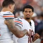 Caleb Williams - Zwei Footballspieler der Chicago Bears stehen während eines Spiels in ihrer Spielkleidung am Spielfeldrand und unterhalten sich. Unter ihnen ist Bears-Quarterback Caleb Williams, der offenbar mit seinem Teamkollegen über die Strategie spricht, nachdem er sich kürzlich eine Verletzung zugezogen hat. Alt-Text wurde KI-generiert.
