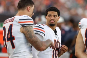 Caleb Williams - Zwei Footballspieler der Chicago Bears stehen während eines Spiels in ihrer Spielkleidung am Spielfeldrand und unterhalten sich. Unter ihnen ist Bears-Quarterback Caleb Williams, der offenbar mit seinem Teamkollegen über die Strategie spricht, nachdem er sich kürzlich eine Verletzung zugezogen hat. Alt-Text wurde KI-generiert.