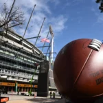 Australien - Melbourne - Vor dem Eingang eines Stadions steht unter blauem Himmel mit vereinzelten Wolken und einem Baum in der Nähe eine große American-Football-Skulptur, die Fans von NFL-Teams und Reisende, die ihre Reisen für 2026 planen, dazu einlädt, die lebhafte Atmosphäre zu genießen. Alt-Text wurde KI-generiert.