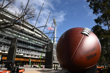 Australien - Melbourne - Vor dem Eingang eines Stadions steht unter blauem Himmel mit vereinzelten Wolken und einem Baum in der Nähe eine große American-Football-Skulptur, die Fans von NFL-Teams und Reisende, die ihre Reisen für 2026 planen, dazu einlädt, die lebhafte Atmosphäre zu genießen. Alt-Text wurde KI-generiert.