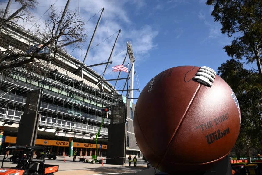 Australien - Melbourne - Vor dem Eingang eines Stadions steht unter blauem Himmel mit vereinzelten Wolken und einem Baum in der Nähe eine große American-Football-Skulptur, die Fans von NFL-Teams und Reisende, die ihre Reisen für 2026 planen, dazu einlädt, die lebhafte Atmosphäre zu genießen. Alt-Text wurde KI-generiert.