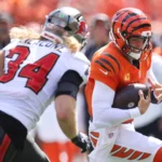 Joe Burrow - Ein Footballspieler in einem orangefarbenen Trikot läuft mit dem Ball, während ein Spieler in einem weißen Trikot mit der Nummer 34 versucht, ihn während eines Spiels zu tackeln, da Burrow, der mit Rückenproblemen zu kämpfen hat, nicht auf dem Spielfeld erwartet wird. Alt-Text wurde KI-generiert.
