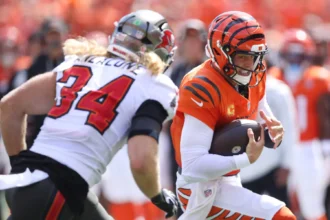 Joe Burrow - Ein Footballspieler in einem orangefarbenen Trikot läuft mit dem Ball, während ein Spieler in einem weißen Trikot mit der Nummer 34 versucht, ihn während eines Spiels zu tackeln, da Burrow, der mit Rückenproblemen zu kämpfen hat, nicht auf dem Spielfeld erwartet wird. Alt-Text wurde KI-generiert.